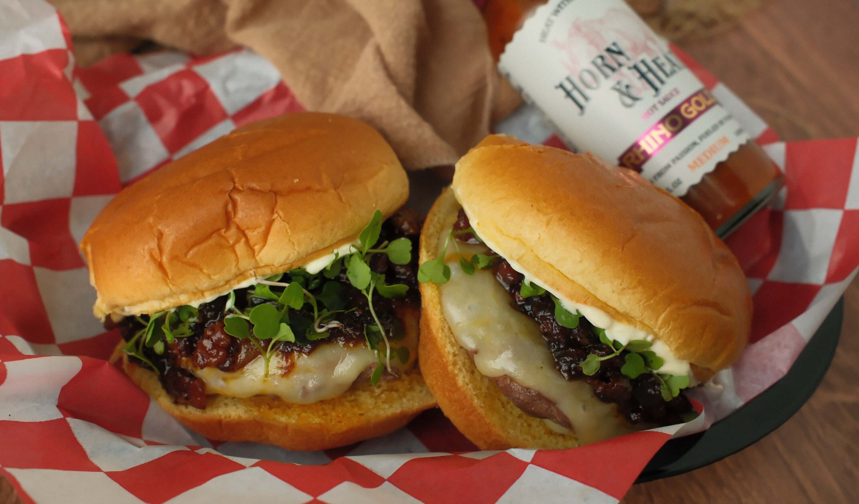 Horn & Heat Burger with Rhino Gold Bacon Jam served on a plate with checkered paper and bottle of sauce in the background.