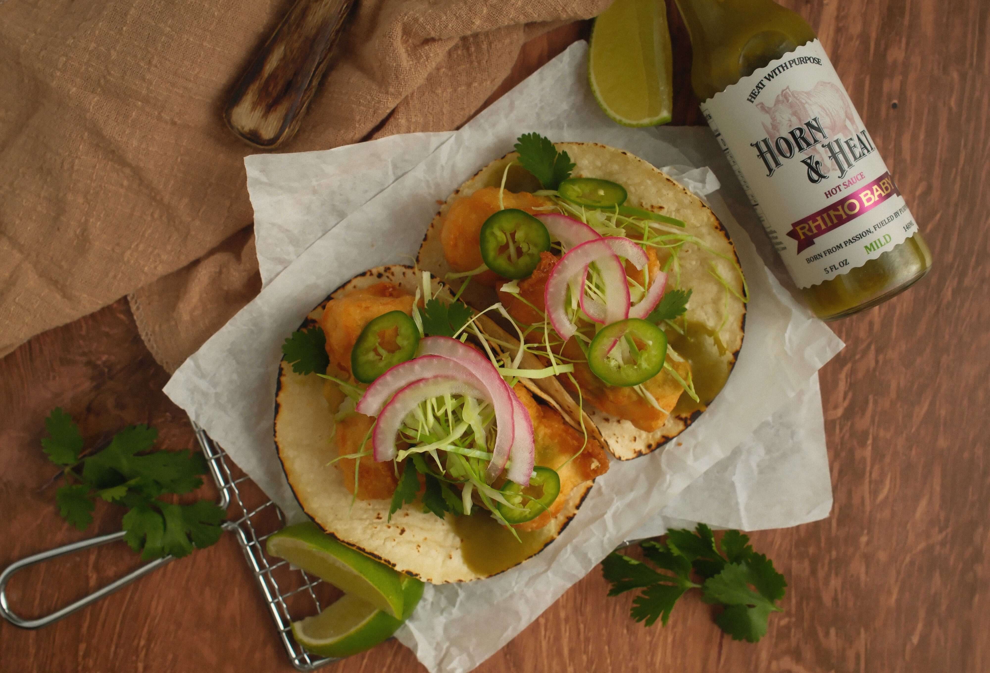 Horn & Heat Tempura Fried Fish Tacos with toppings and a bottle of sauce in the background.